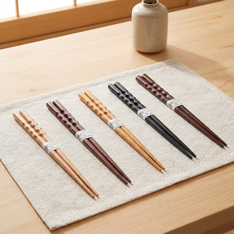 Five pairs of bamboo Japanese chopsticks with carved grip detail in natural, burgundy, blonde, black and dark brown laid on a Japanese wave patterned placemat with a ceramic vase in the background