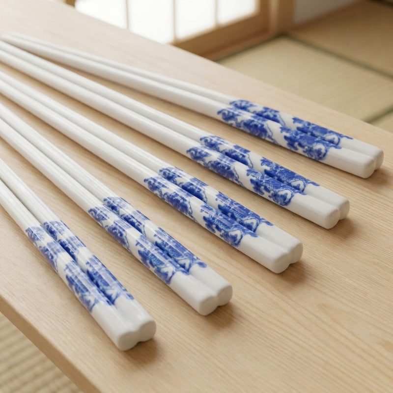 Five pairs of white ceramic chopsticks with flower blue and white porcelain landscape motif fanned out on a light wood surface near a shoji window