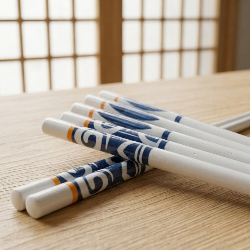 Close-up of white ceramic chopsticks with blue indigo motifs and orange accent bands resting on a wood surface with a shoji screen in the background