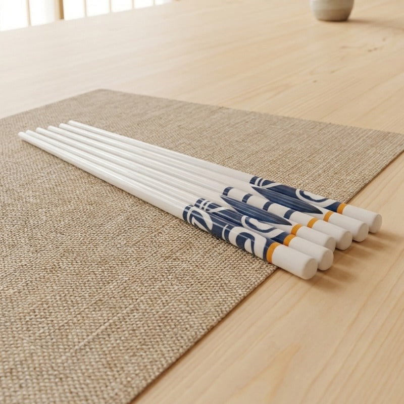 Five pairs of white ceramic chopsticks with blue wave patterns and orange accent bands laid on a woven linen placemat on a light wood table