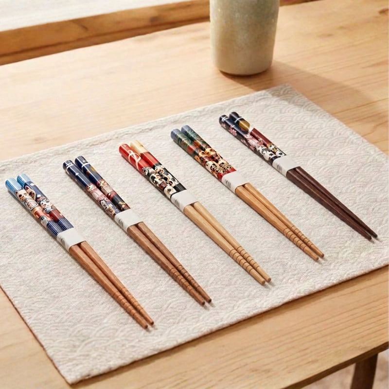 Five pairs of wood chopsticks with colorful painted designs lie diagonally across a woven placemat on a wooden table