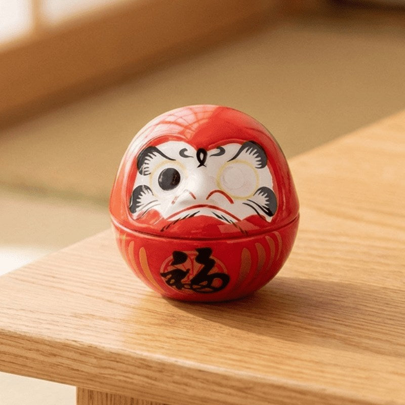 Traditional daruma art in vivid red with determined facial expression displayed on wooden surface catching soft natural daylight