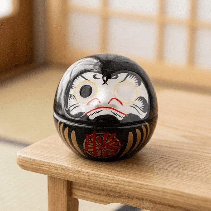 Lacquered daruma black with gold and red decorative elements sitting on wooden table beside bright window in minimalist Japanese interior