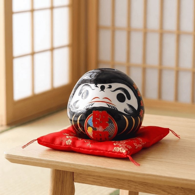 Traditional daruma doll black japanese ceramic with fierce expression and colorful details resting on red silk cushion in peaceful tatami room setting