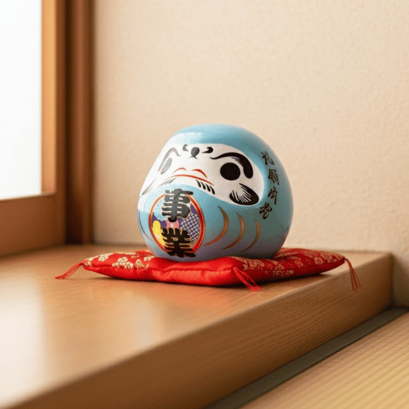 Career focused daruma doll blue with calligraphy details sitting on decorative red cushion near traditional shoji screen with warm natural lighting