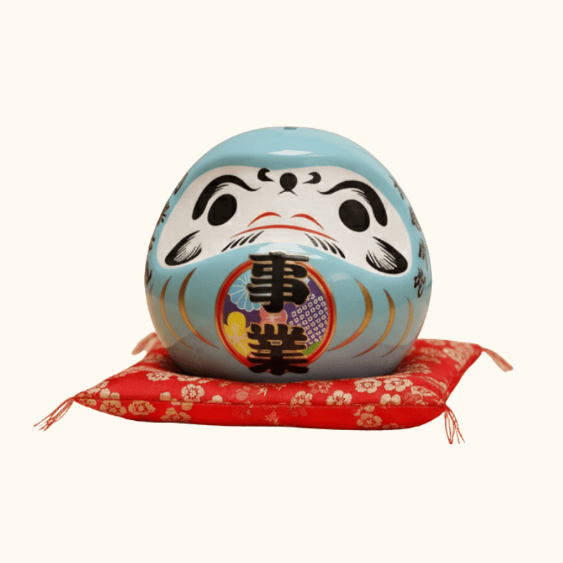 Professional daruma doll japanese blue with colorful kanji belly displayed on red floral cushion against clean cream background for clear detailed view