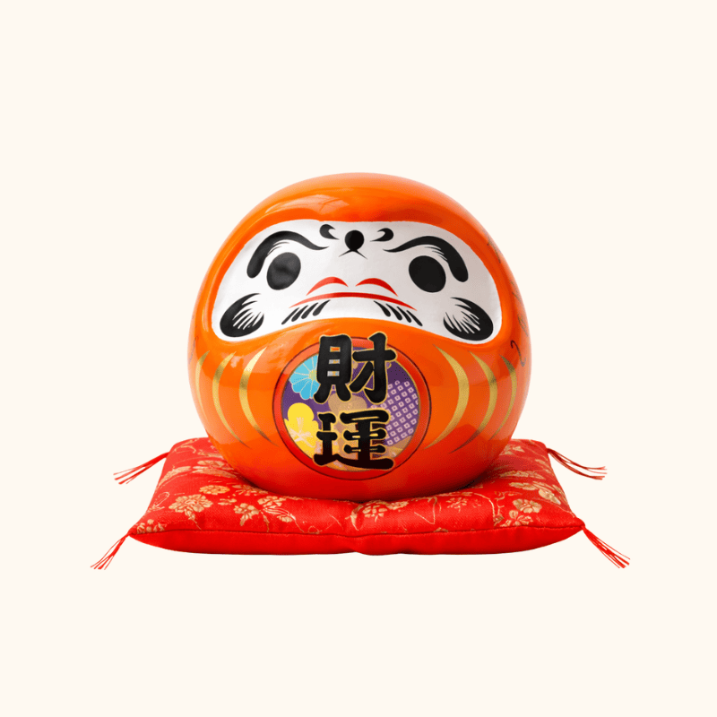 Entrepreneurial daruma japanese ceramic in glossy orange finish with colorful kanji details displayed on red floral cushion against clean white background