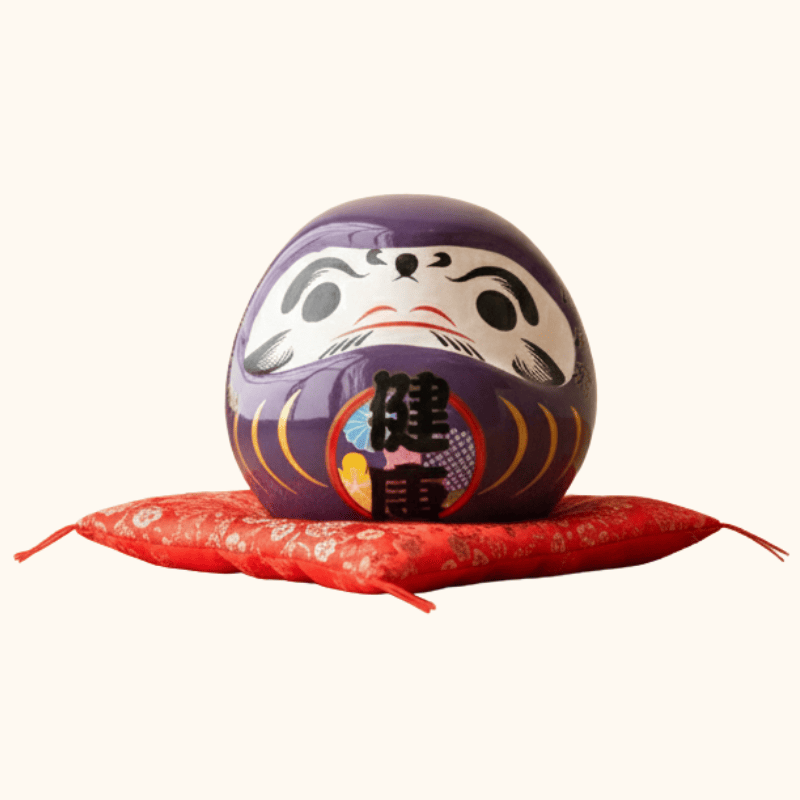 Health symbol kanji on daruma japanese doll purple in rich purple finish with traditional calligraphy displayed on red floral cushion against clean white backdrop
