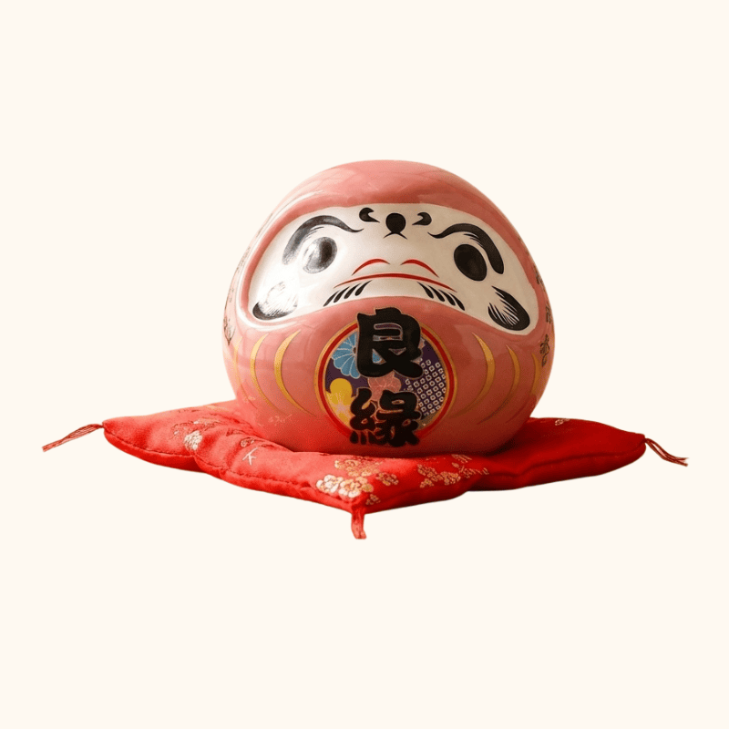 Romantic daruma japanese pink in soft rose ceramic with traditional kanji details displayed on red floral cushion against clean white background view
