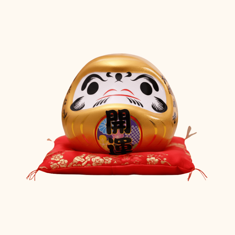 Wealth intention gold japanese daruma in metallic ceramic with prosperity kanji displayed on red floral cushion against clean white background view