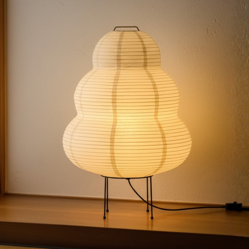 Japanese paper floor lamp with ribbed washi gourd shaped shade on black metal tripod glowing warm light on wooden shelf surface