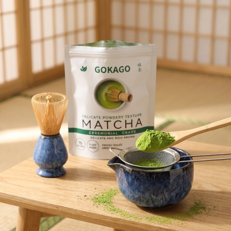 Vibrant green Japanese matcha powder held above a fine mesh sieve, with a ceramic tea bowl, bamboo whisk and GOKAGO packaging.