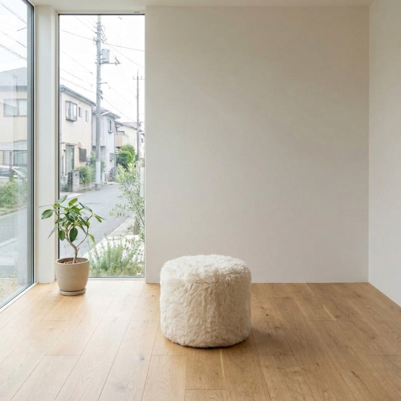 Low Japanese sofa pouf in cream shaggy fur positioned in a minimalist room with floor to ceiling window overlooking a residential street and a potted olive tree