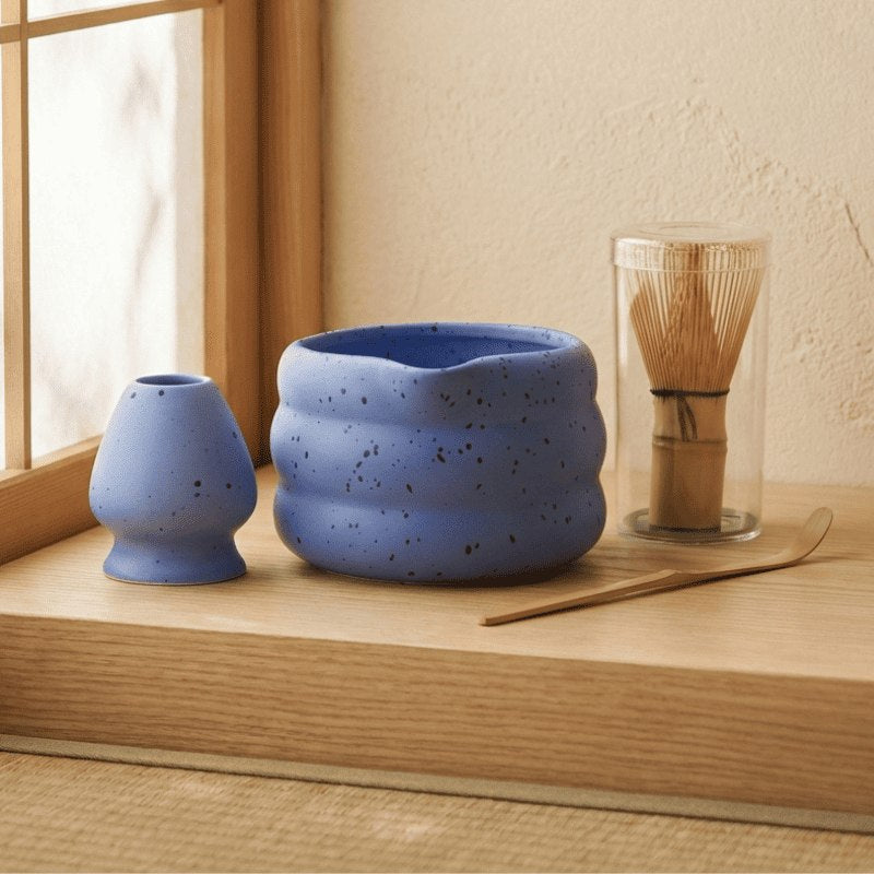 Blue speckled ceramic matcha tea whisk set with bamboo scoop and whisk holder arranged on wooden table near bright window in minimalist setting
