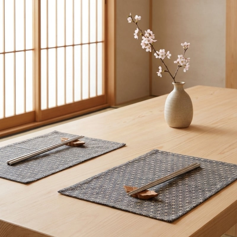 Two place settings with metal Japanese chopsticks in stainless steel on leaf-shaped wooden rests laid on dark geometric patterned placemats, ceramic vase with cherry blossom branches and shoji door in the background