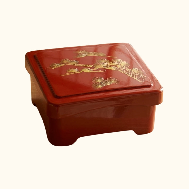 Crimson red sushi bento box with decorative golden pine branches etched on lid against clean white background