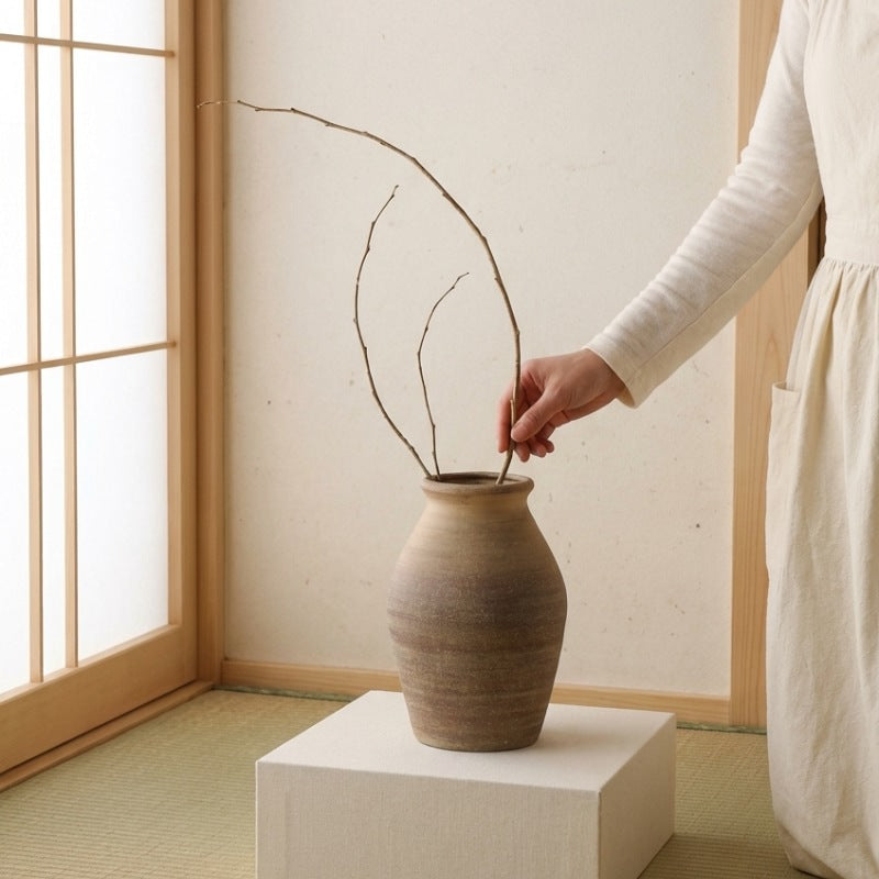 Hand arranging curved branches in rustic vase ikebana ceramic on fabric covered pedestal over tatami