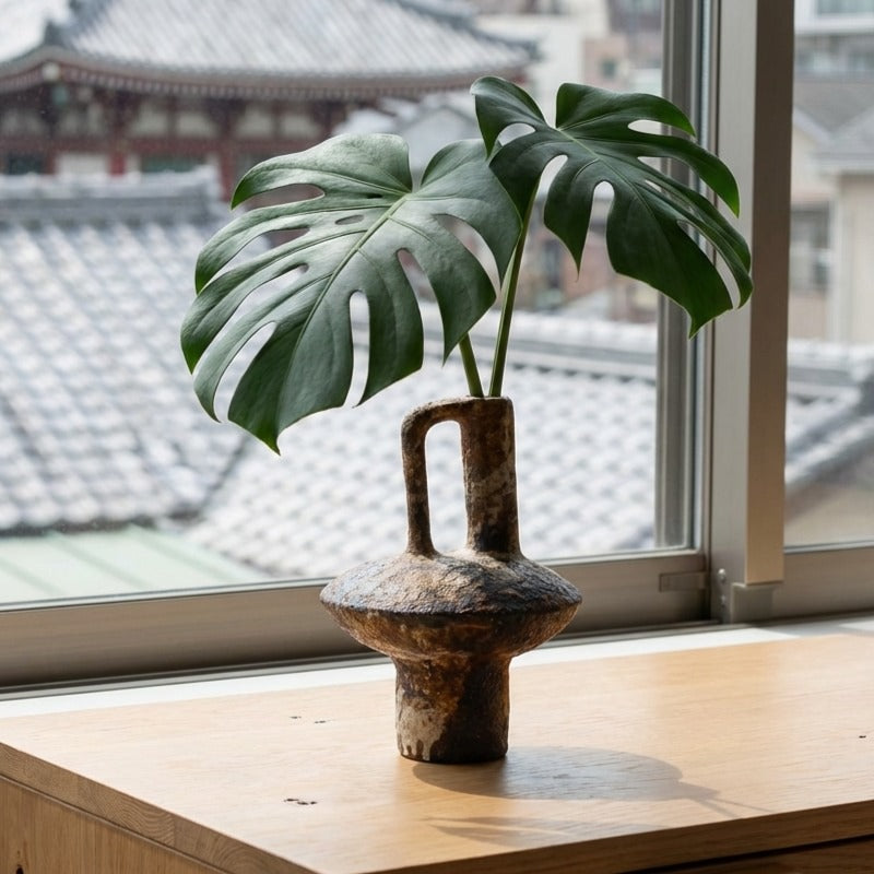 Sculptural wabi sabi vase holding fresh monstera leaf by window overlooking traditional japanese temple rooftops
