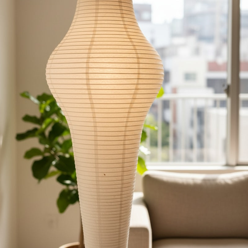 Tall akari lamp paper with elegant hourglass silhouette rice paper shade beside modern sofa near bright apartment window with city views
