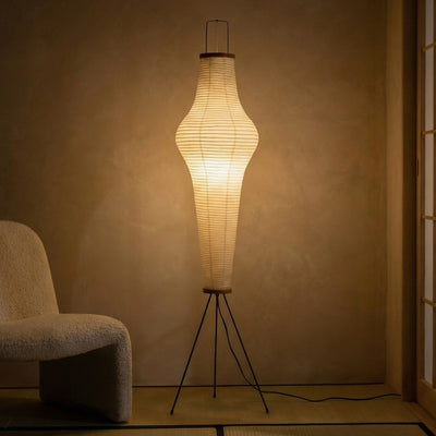 Glowing akari lamp illuminating traditional tatami room with sculptural rice paper cylinder shade on black tripod beside cream armchair at night