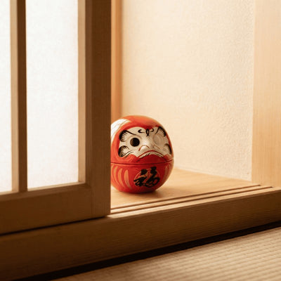 Vibrant red art daruma with bold brushstroke design sitting on wooden table near traditional window in warm ambient Japanese interior