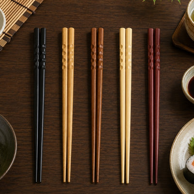 Five pairs of bamboo chopsticks in black, natural, brown, cream and dark red laid flat on a dark wood table next to sushi rolls, soy sauce and a bamboo mat