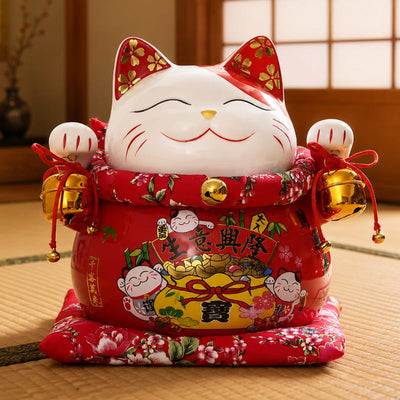Smiling big maneki neko figurine with gold-flecked ears and jingling bells, seated on a red floral cushion on a wooden table with tatami flooring and a shoji screen window behind it