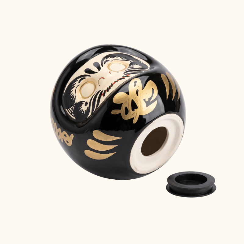 Underside of a black Japanese Daruma money box, showing its removable stopper and traditional gold decorations.