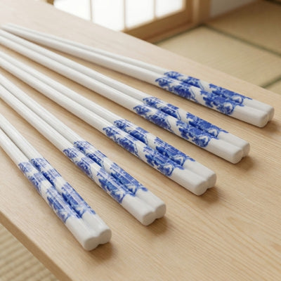 Five pairs of white ceramic chopsticks with flower blue and white porcelain landscape motif fanned out on a light wood surface near a shoji window