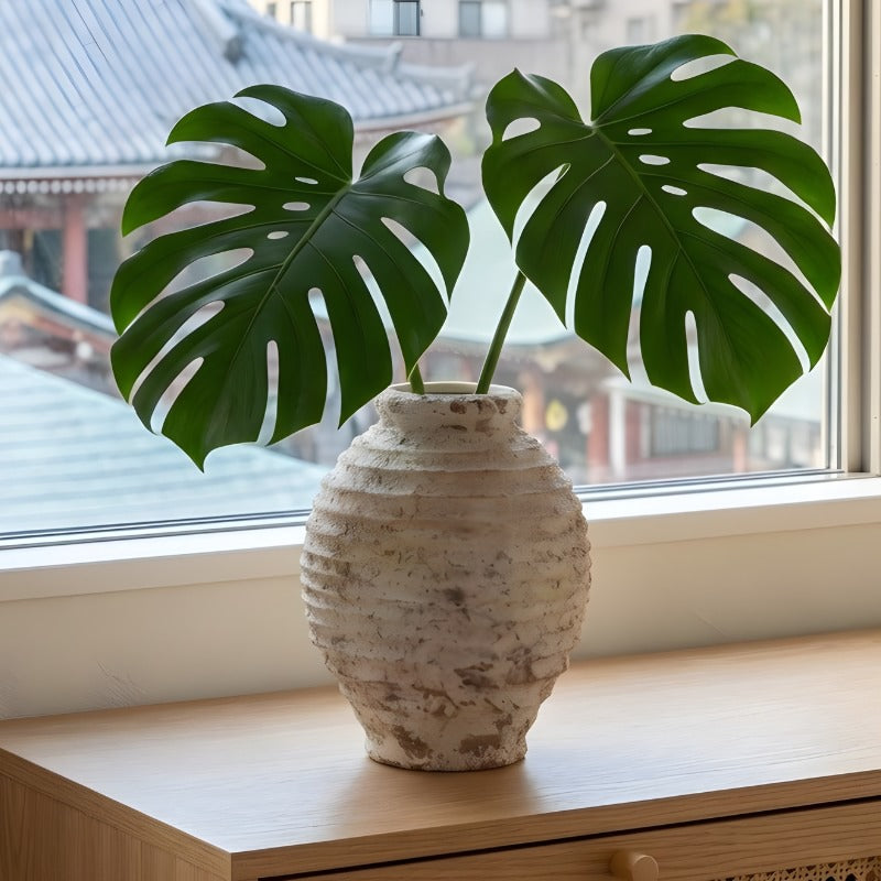 Sculptural ceramic ikebana vase with ribbed texture holding monstera leaves on windowsill with traditional japanese architecture view