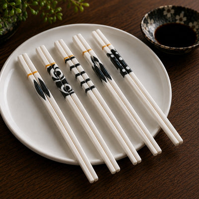 Five pairs of white ceramic chopsticks with black geometric patterns and gold accents arranged on a round white plate, soy sauce bowl visible on dark wood table