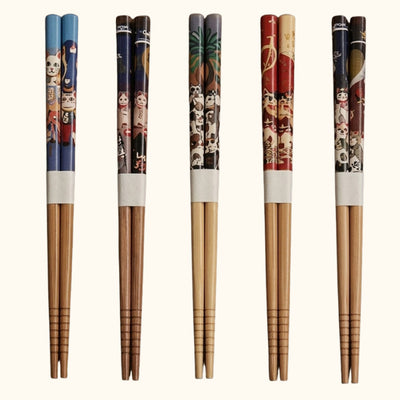 Five pairs of kawaii wood chopsticks with playful cat and character designs stand upright against a cream background