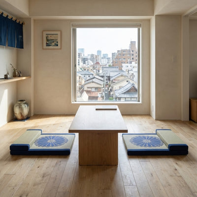 Cushion blue for meditation pair with chrysanthemum patterns flanking wooden table near window overlooking traditional urban rooftops with noren
