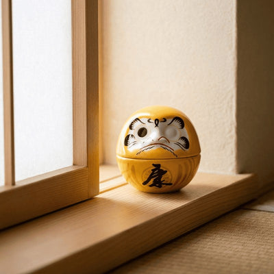 Classic daruma antique in golden yellow with determined expression sitting on wooden table beside traditional window in soft ambient light