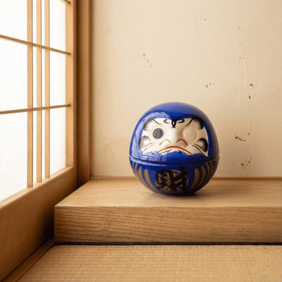 Traditional daruma blue figurine with blank eyes and gold accents resting on wooden surface near shoji window in minimalist setting