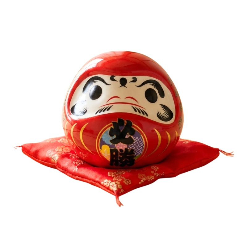Lucky daruma doll japanese red with golden kanji details displayed on red silk cushion against clean cream background showing traditional Japan art