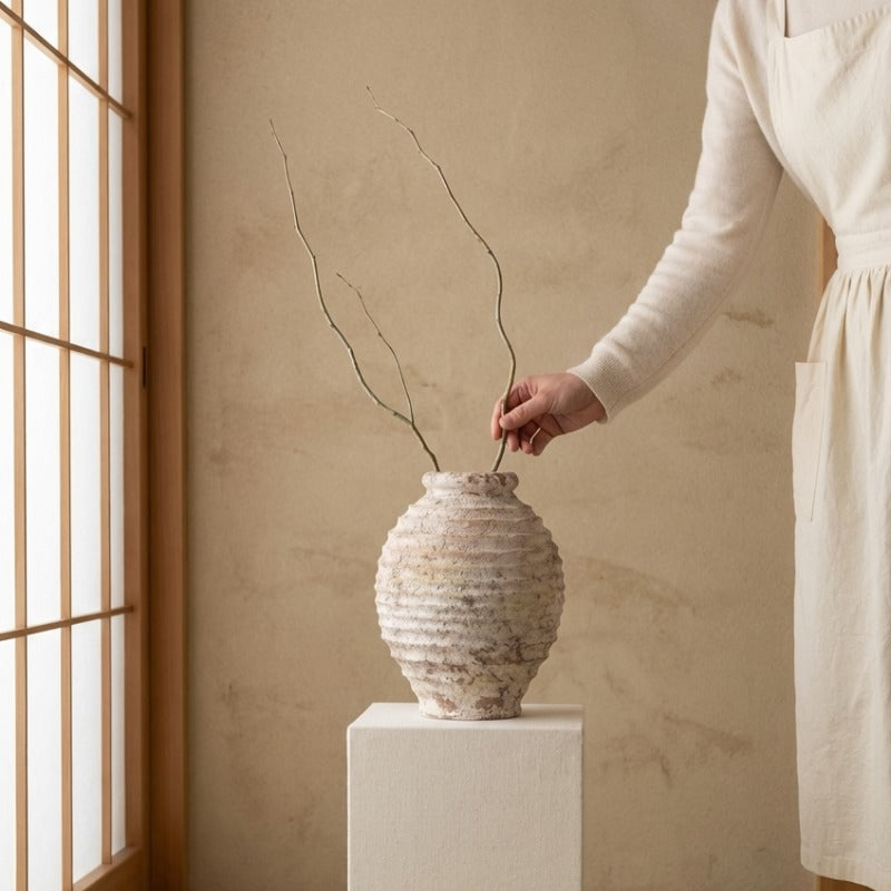Hand placing delicate branches in organic ikebana ceramic vase with horizontal ridges on fabric pedestal