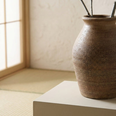 Minimalist ikebana vase brown with textured surface holding branches on white pedestal near shoji screen