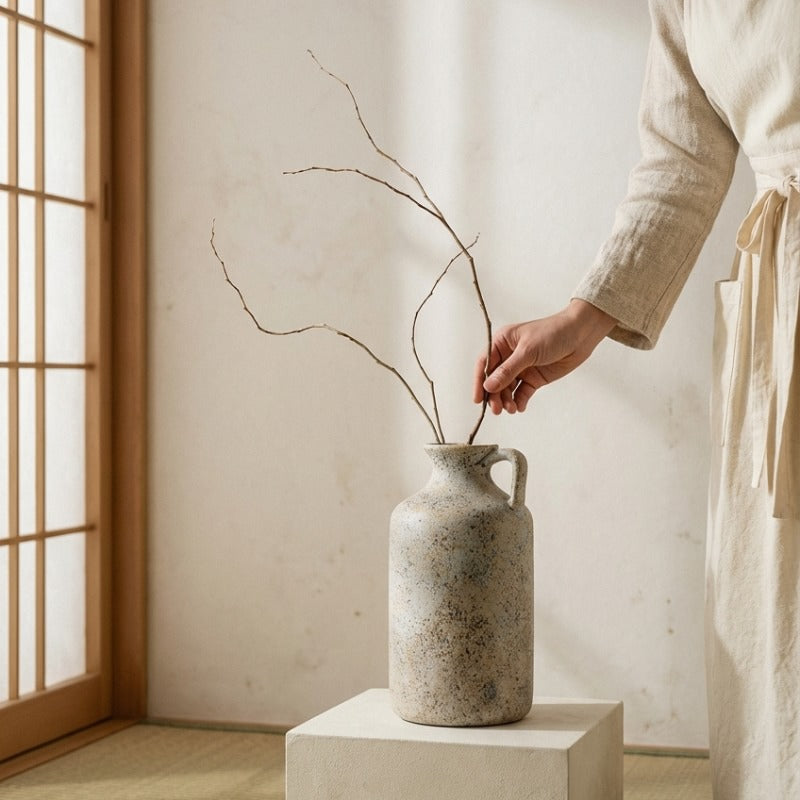Hand arranging minimalist branches in rustic ikebana vintage vase with handle on white pedestal near shoji screen