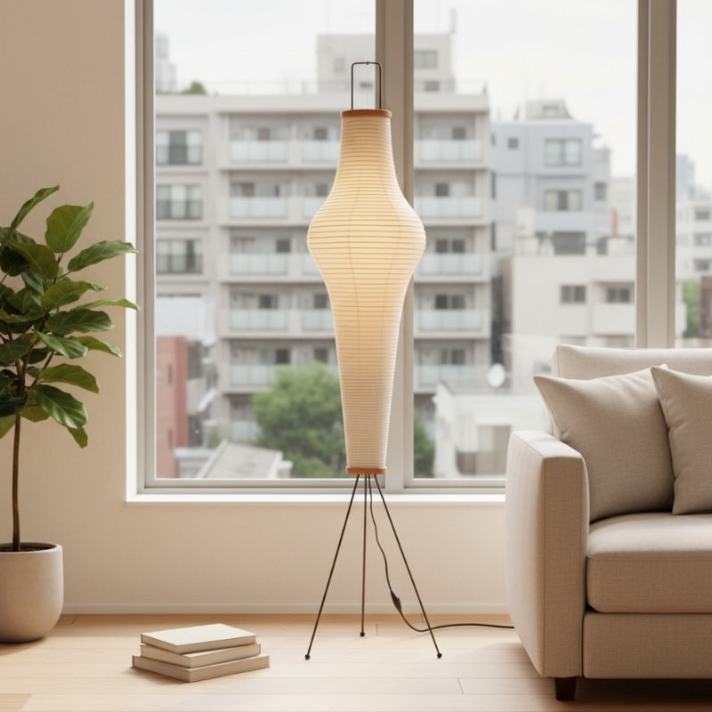 Sculptural japanese akari lamp with elongated rice paper cylinder glowing beside beige sofa near large window overlooking urban neighborhood