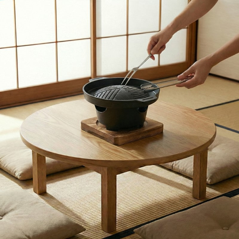Japanese barbecue style cooking preparation with metal tongs over round ridged grill on low table with floor cushions