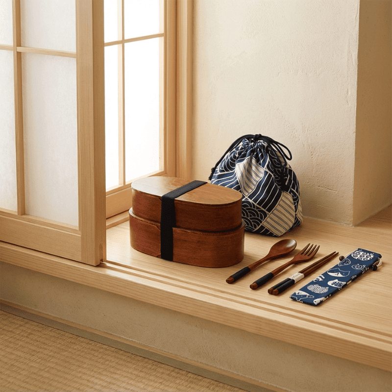 Two-tier dark wood japanese bento box traditional with black elastic band displayed on windowsill with patterned fabric carrier bag and wooden utensils in warm natural light