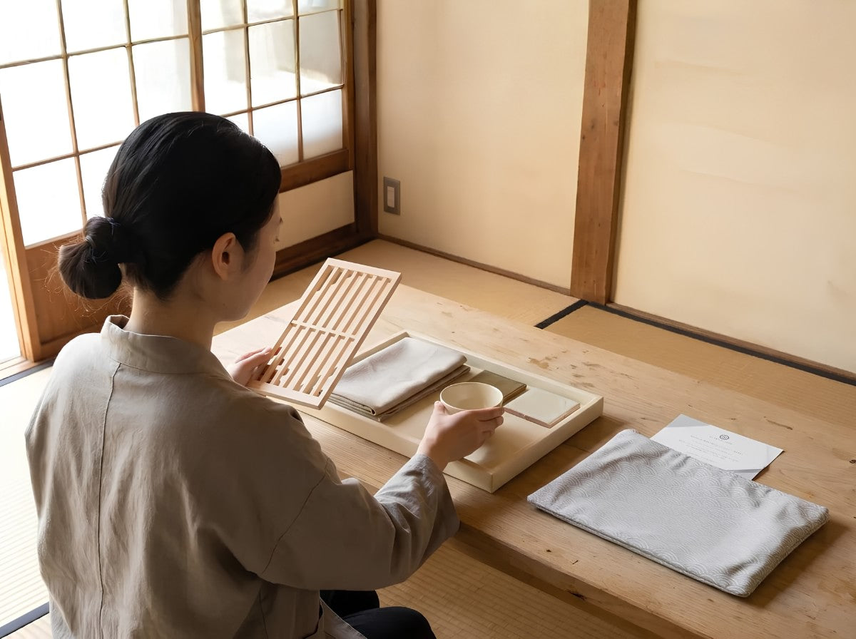 A woman in a beige linen shirt sits on a tatami floor in a traditional Japanese room, holding a small wooden cup and demonstrating the use of a tea tray set with a folded gray cloth pouch and white paper on a low wooden table.