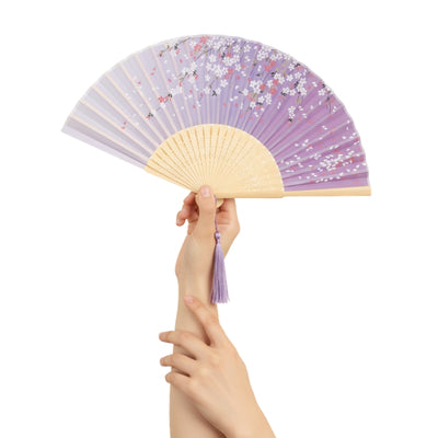 Hand holding japanese folding fan paper in soft lavender with delicate sakura blossoms carved bamboo ribs and matching purple tassel