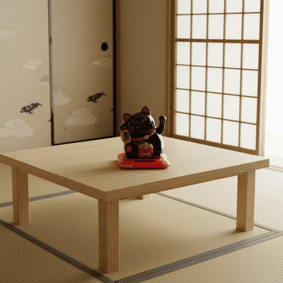 Traditional japanese maneki neko black cat figurine with waving paw on low wooden table beside shoji screen and bird art panel in tatami room