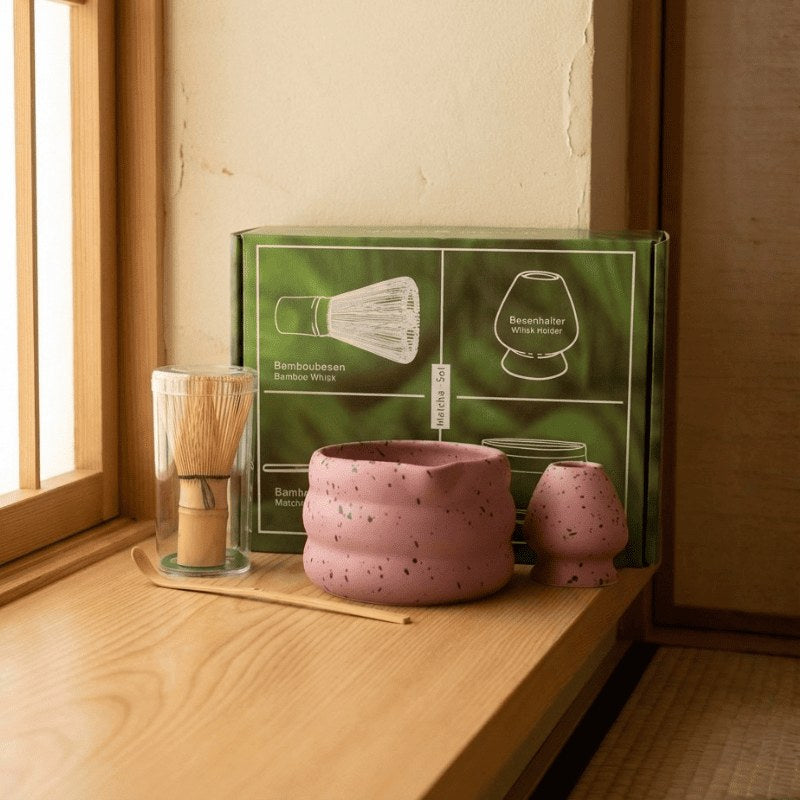 Pink ceramic matcha gift set with bamboo whisk and green illustrated box displayed on wooden shelf near bright window in traditional interior