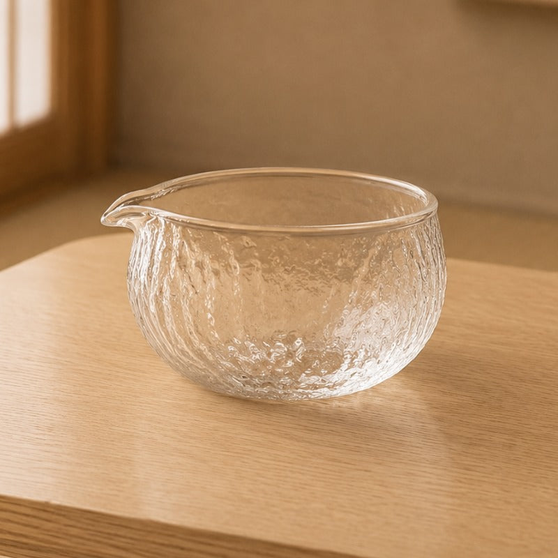 Handblown clear glass Japanese matcha tea bowl with hammered texture and small pouring spout on a light wood table, shoji window in background