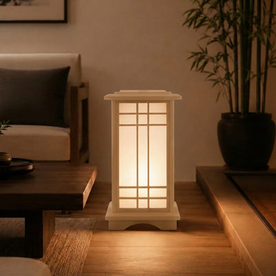 Illuminated shoji-style japanese lamp in a small lantern design glowing on a wooden step beside a dark coffee table, potted bamboo, and neutral sofa in a cozy living room