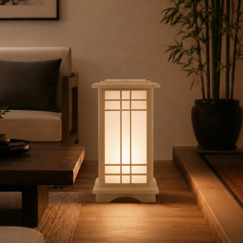 Illuminated shoji-style japanese lamp in a small lantern design glowing on a wooden step beside a dark coffee table, potted bamboo, and neutral sofa in a cozy living room