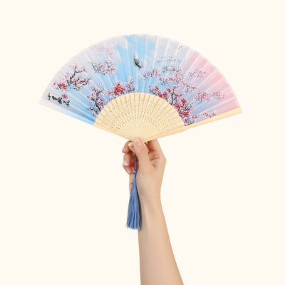 Hand holding japanese vintage hand fan with watercolor sakura pattern in soft blue and pink tones with blue tassel against white background
